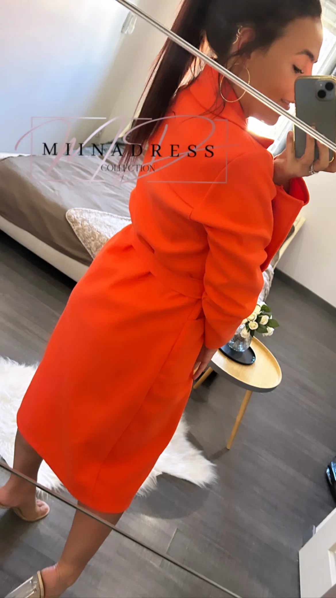 Femme portant un manteau orange ceinturé, look chic, dans une chambre moderne.