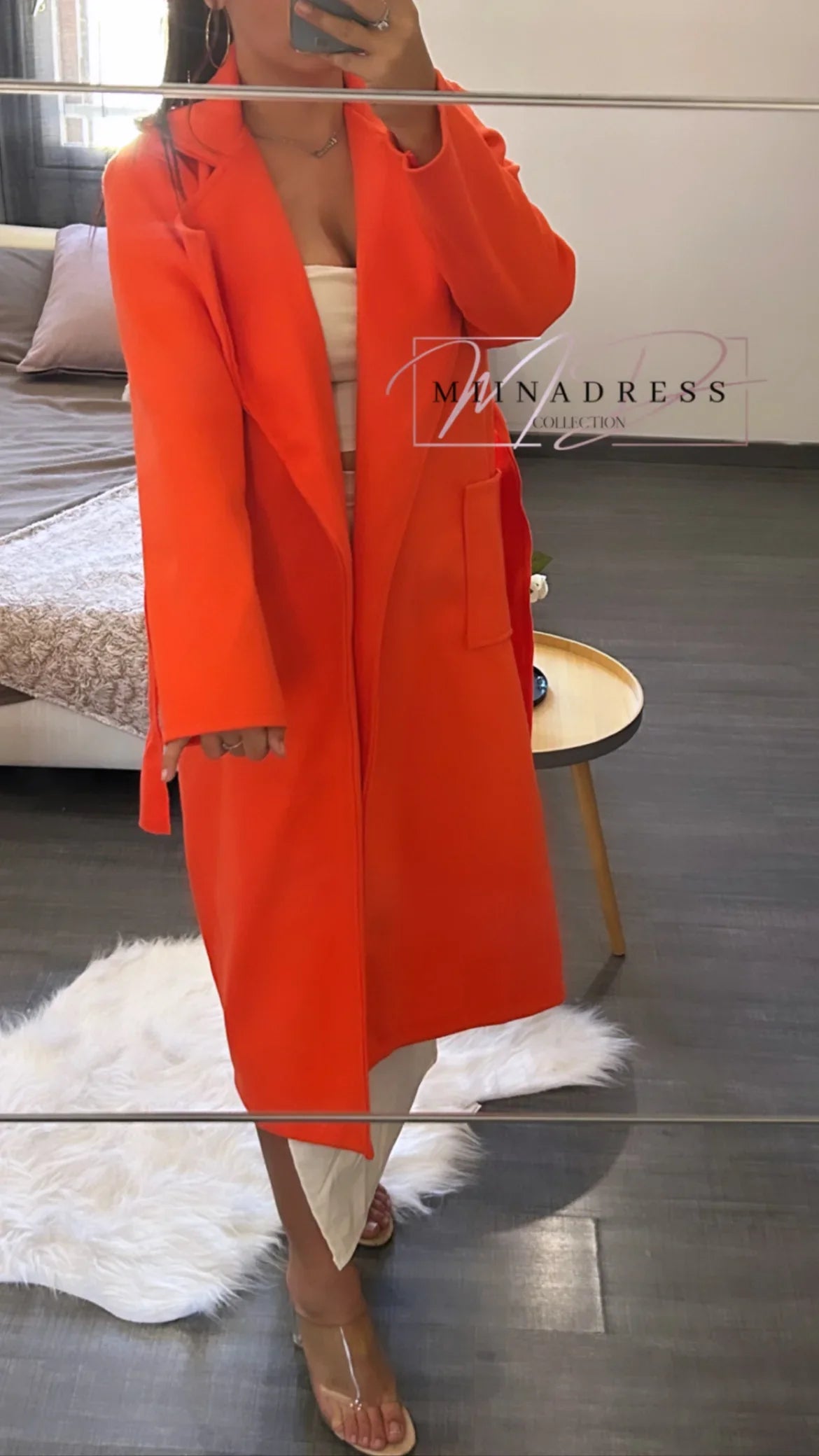 Femme portant un manteau long orange chic, debout dans une chambre moderne