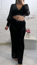 Femme portant un ensemble noir brillant, crop top à manches longues et pantalon chic, devant un mur blanc.