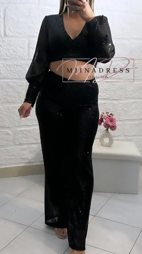 Femme portant un ensemble noir brillant, crop top à manches longues et pantalon chic, devant un mur blanc.