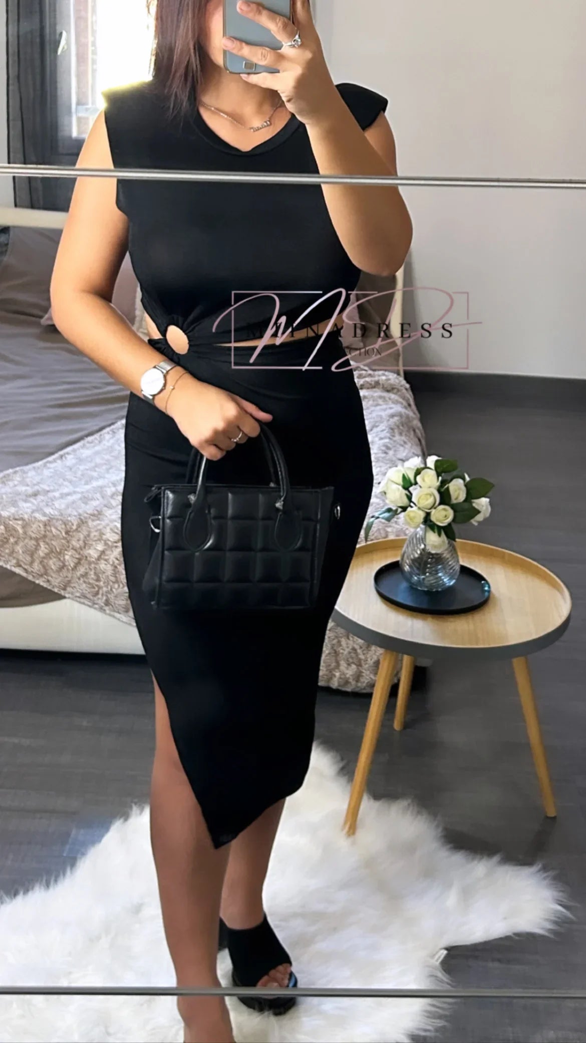 Femme en robe noire chic, sac matelassé, posant dans une chambre moderne, style élégant.
