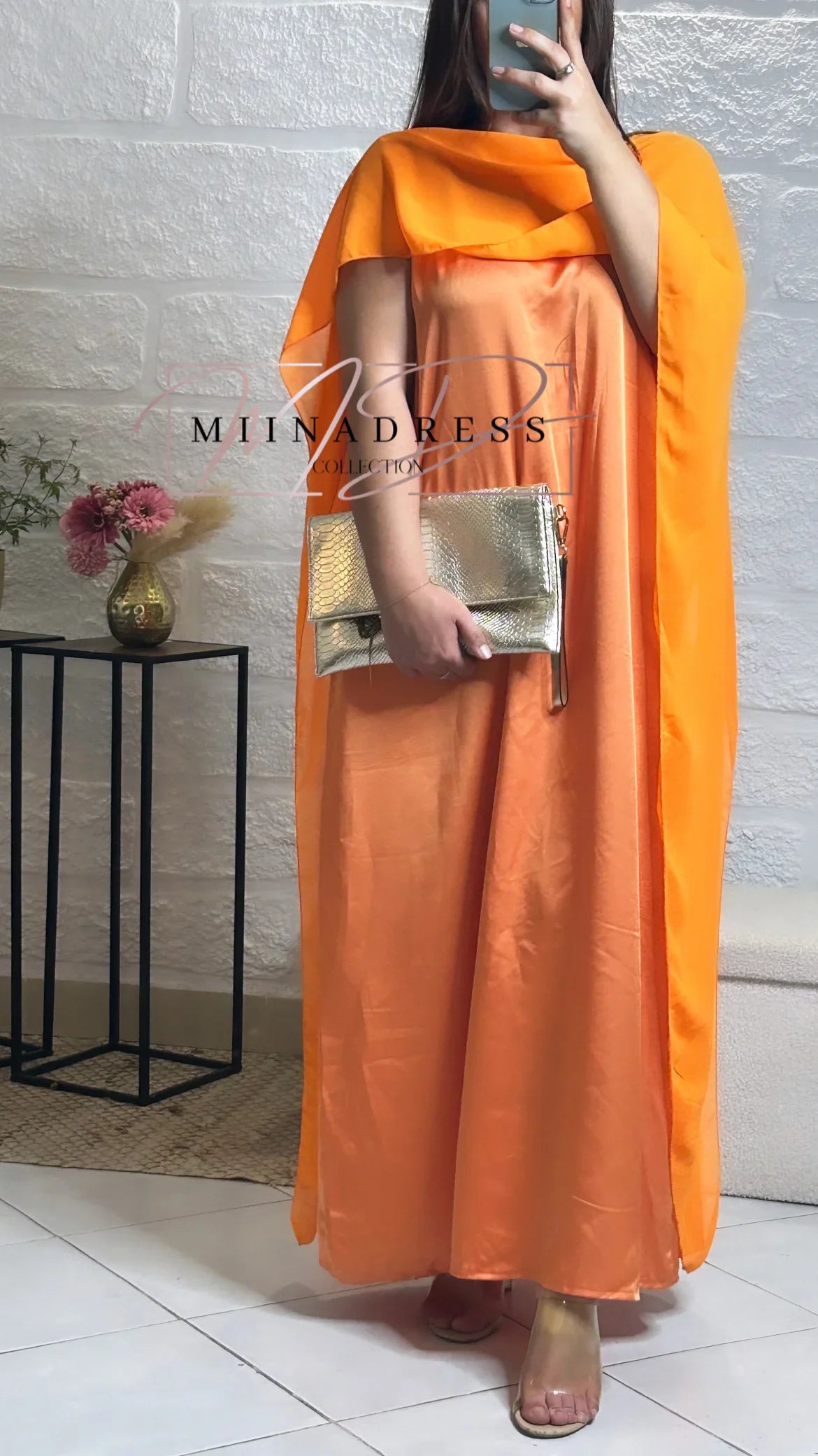 Femme portant une robe longue orange satinée, pochette dorée, déco florale en fond