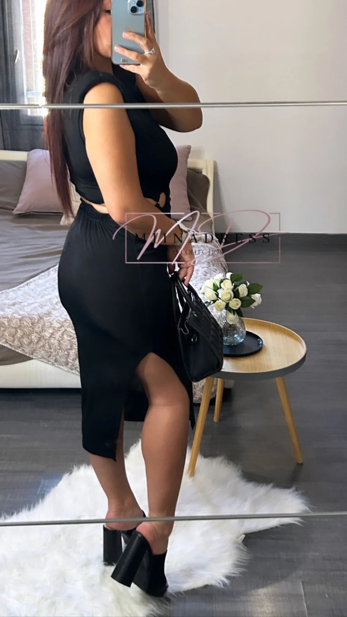 Femme en robe noire fendue, sac à main et talons, posant dans une chambre moderne.