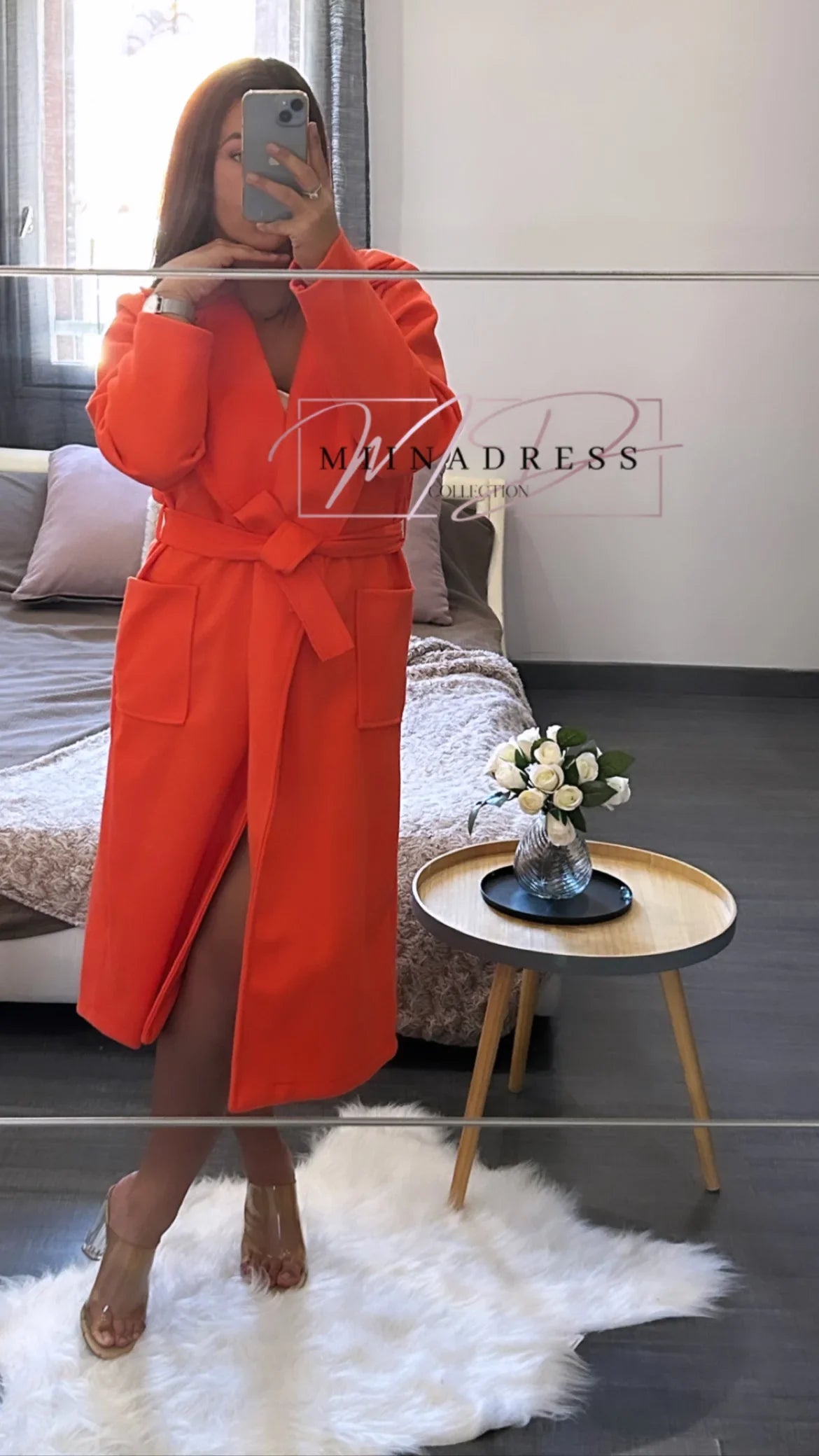 Femme en manteau long orange ceinturé, devant miroir, déco chambre moderne, fleurs blanches.