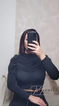Femme portant un top noir à manches longues froncé, selfie devant un mur géométrique.