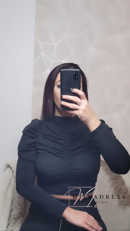Femme portant un top noir à manches longues froncé, selfie devant un mur géométrique.