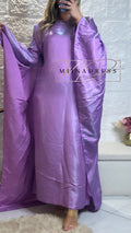 Femme portant une robe longue en satin violet, manches larges, sur fond mural blanc.