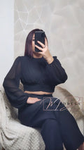 Femme portant un crop top noir manches longues et pantalon large, assise sur un fauteuil cosy.