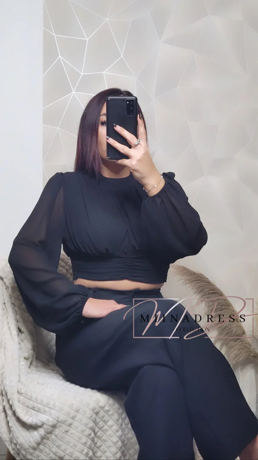Femme portant un crop top noir manches longues et pantalon large, assise sur un fauteuil cosy.